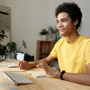 Teenager smiling while studying online at home. Modern education setup with computer and VR headset.